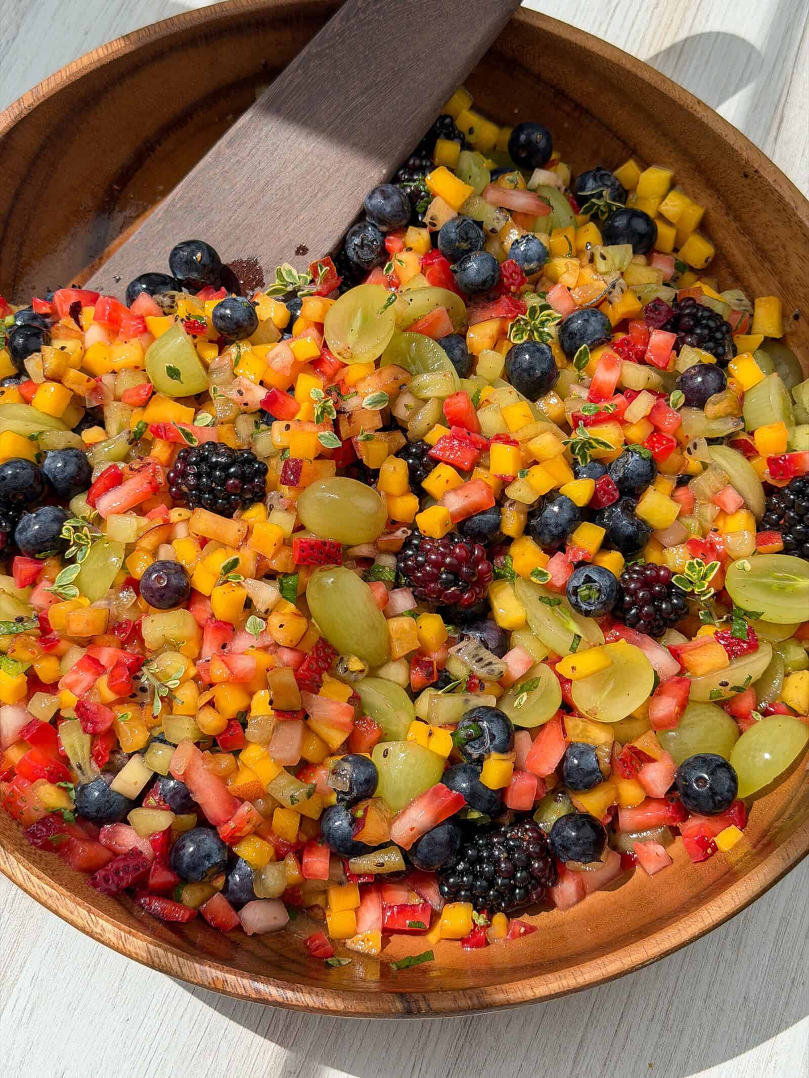 A wooden bowl filled with a colorful fruit salad featuring diced mango, watermelon, strawberries, green grapes, blueberries, blackberries, and a few small green herbs, with a wooden serving spoon.