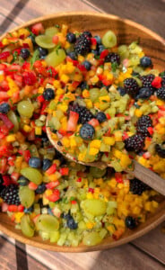 A wooden bowl filled with a colorful fruit salad, including diced mango, kiwi, strawberries, grapes, blueberries, and blackberries. A wooden spoon scoops up a portion of the vibrant mix.