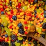 A wooden spoon holds a colorful mix of diced fruit, including mango, red berries, green grapes, blueberries, and blackberries, over a large bowl filled with the same vibrant fruit salad.
