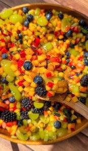 A wooden spoon holds a colorful mix of diced fruit, including mango, red berries, green grapes, blueberries, and blackberries, over a large bowl filled with the same vibrant fruit salad.