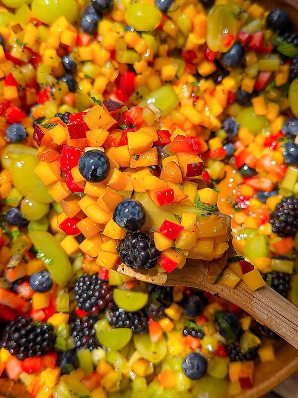 A wooden spoon holds a colorful mix of diced fruit, including mango, red berries, green grapes, blueberries, and blackberries, over a large bowl filled with the same vibrant fruit salad.