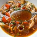 A close-up of No Water Chicken Soup with a spoonful of clear brown broth held above it, showing pieces of chicken, chopped carrots, and fresh herbs on top—perfect for a rich, waterless chicken soup recipe.