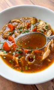 A close-up of No Water Chicken Soup with a spoonful of clear brown broth held above it, showing pieces of chicken, chopped carrots, and fresh herbs on top—perfect for a rich, waterless chicken soup recipe.