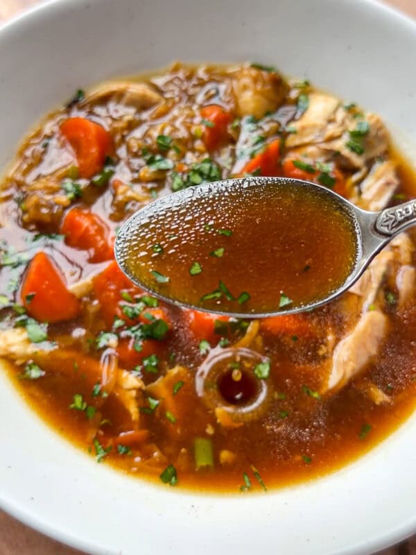 A close-up of No Water Chicken Soup with a spoonful of clear brown broth held above it, showing pieces of chicken, chopped carrots, and fresh herbs on top—perfect for a rich, waterless chicken soup recipe.