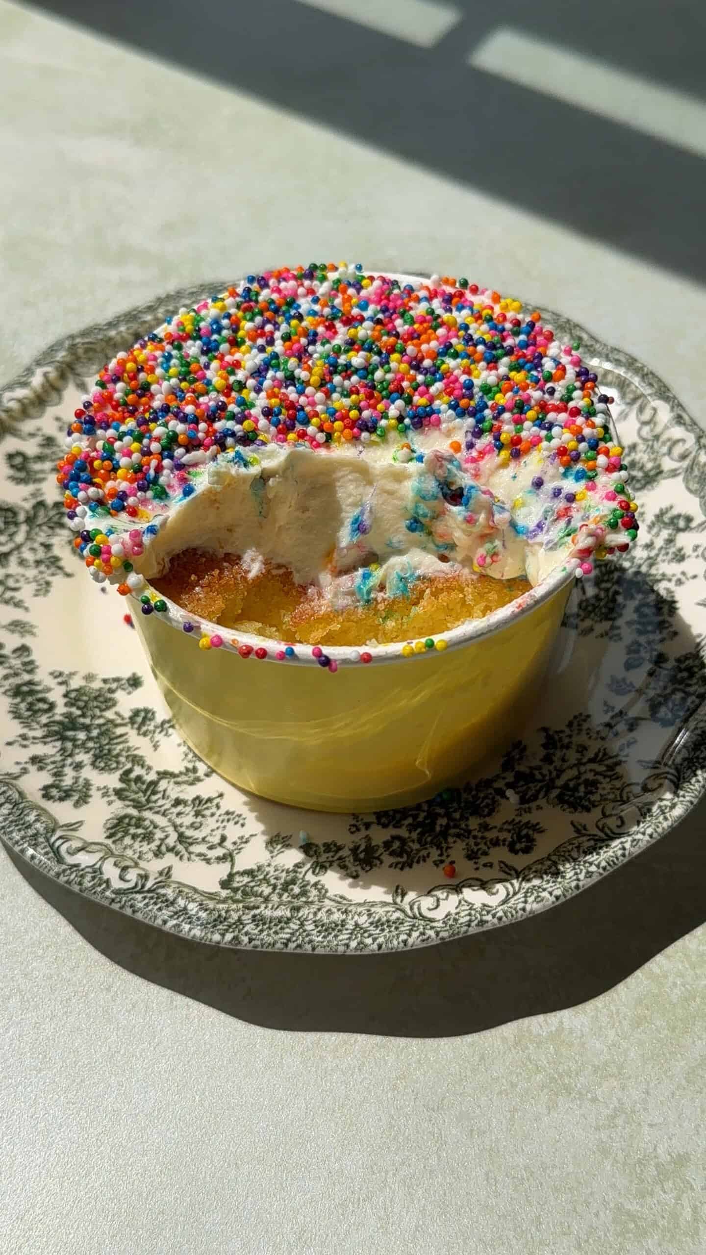A small round Viral Dot Cake in a yellow cup, topped with white frosting and colorful sprinkles, sits on a patterned plate. A portion has been eaten, revealing the cake and frosting layers inside. Sunlight casts shadows on the table.