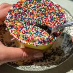 A person holds a cup of ice cream covered in colorful round sprinkles, resembling the look of a Viral Dot Cake, set on a decorative black and white plate. A spoon scoops into the sprinkled ice cream under bright natural light.