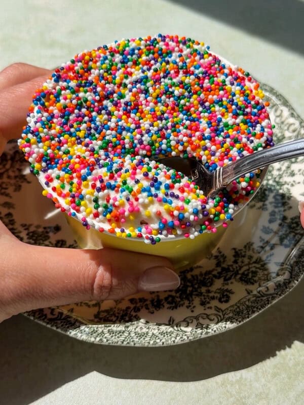 A person holds a cup of ice cream covered in colorful round sprinkles, resembling the look of a Viral Dot Cake, set on a decorative black and white plate. A spoon scoops into the sprinkled ice cream under bright natural light.