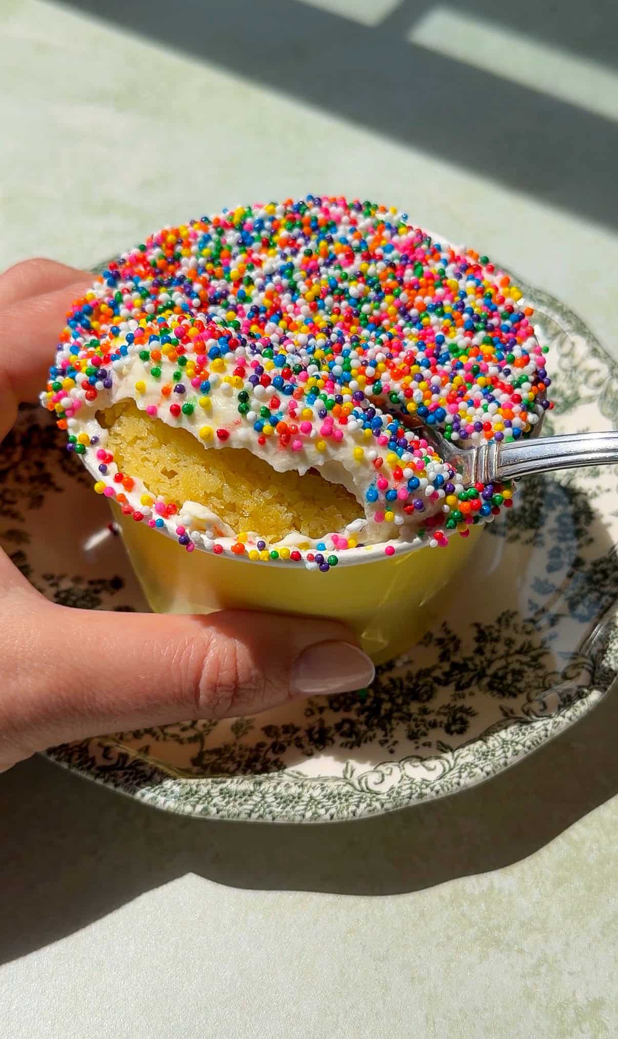 A hand holds a yellow cup filled with Viral Dot Cake, topped with white frosting and colorful rainbow sprinkles. A spoon scoops out a bite, revealing the soft yellow cake inside. The cup sits on a decorative floral-patterned saucer.
