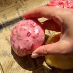 A hand holding a round, pink, textured dessert with red specks rests on brown printed parchment paper in bright sunlight—reminiscent of the vibrant hues in a No Water Chicken Soup recipe. Another yellow item is partially visible nearby.