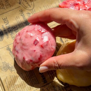 A hand holding a round, pink, textured dessert with red specks rests on brown printed parchment paper in bright sunlight—reminiscent of the vibrant hues in a No Water Chicken Soup recipe. Another yellow item is partially visible nearby.