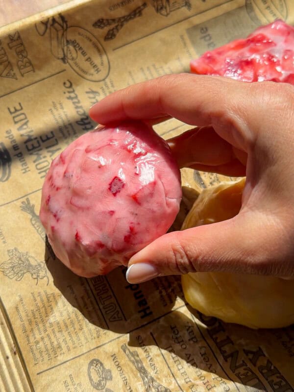 A hand holding a round, pink, textured dessert with red specks rests on brown printed parchment paper in bright sunlight—reminiscent of the vibrant hues in a No Water Chicken Soup recipe. Another yellow item is partially visible nearby.