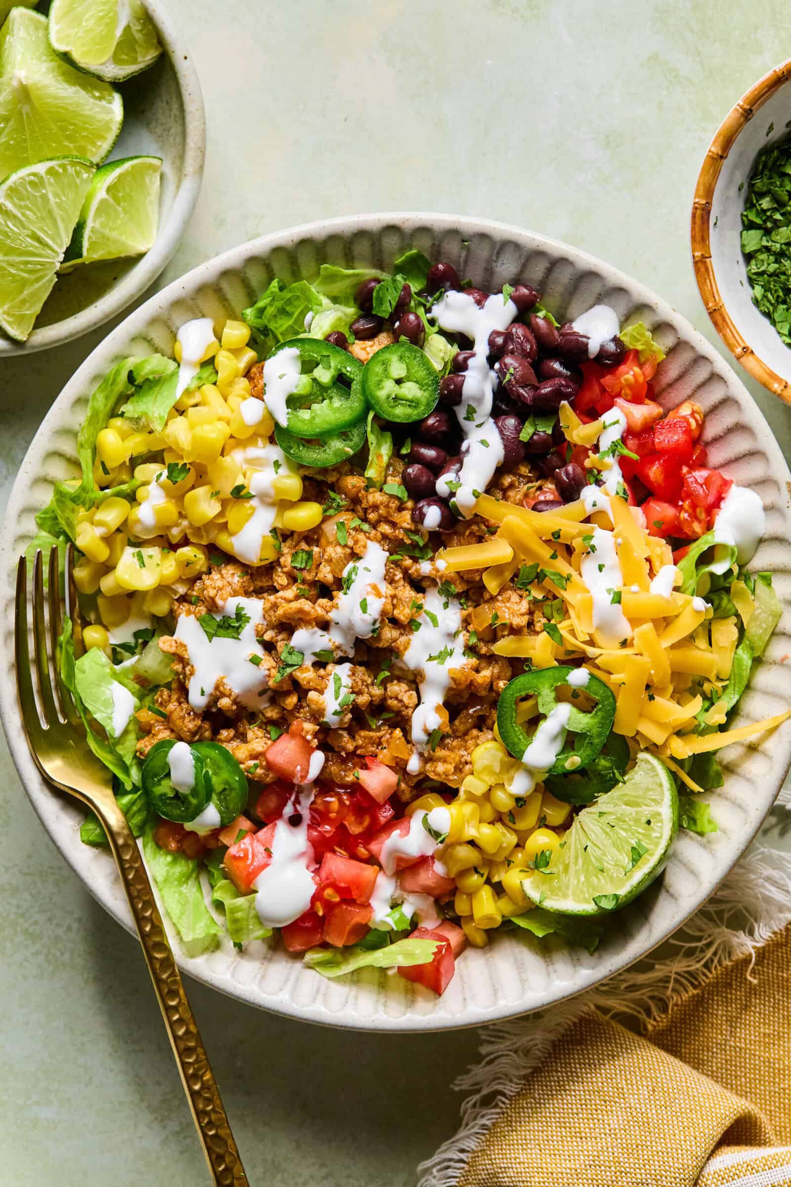 A taco salad in a bowl with ground meat, shredded cheese, black beans, corn, lettuce, tomatoes, jalape&ntilde;os, and a drizzle of white dressing, garnished with lime wedges. A fork, napkin, and a bowl of lime slices are nearby.