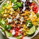 A colorful taco salad in a bowl with lettuce, seasoned ground meat, black beans, corn, diced tomatoes, shredded cheese, jalapeño slices, a lime wedge, and drizzled with white dressing. A gold fork rests on the side.