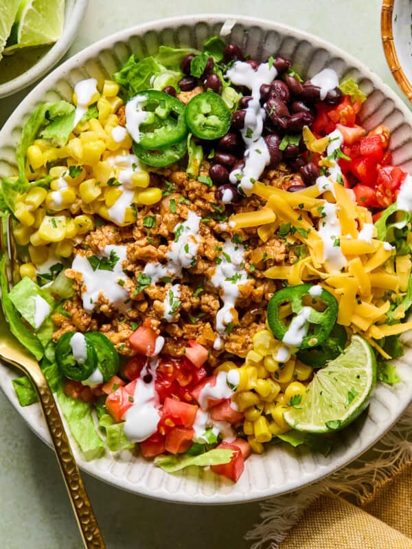 A colorful taco salad in a bowl with lettuce, seasoned ground meat, black beans, corn, diced tomatoes, shredded cheese, jalape&ntilde;o slices, a lime wedge, and drizzled with white dressing. A gold fork rests on the side.