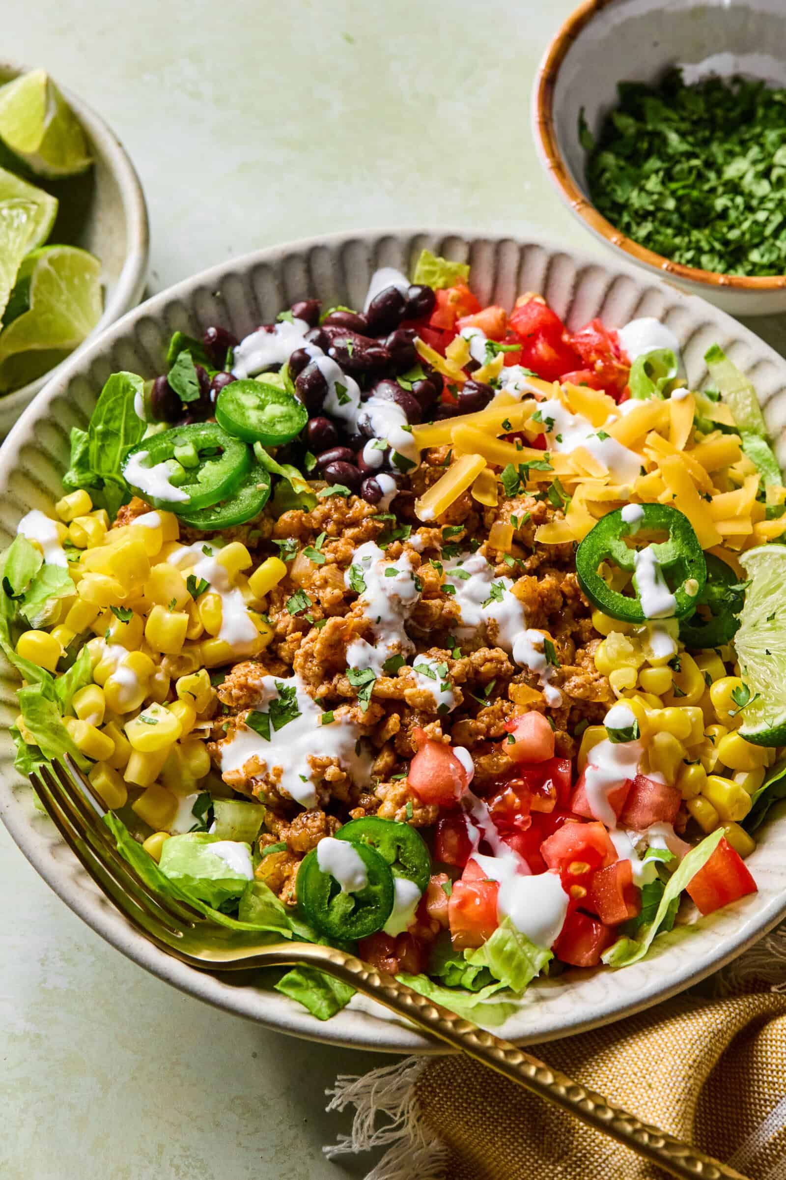 A bowl filled with colorful taco salad ingredients, including lettuce, corn, tomatoes, black beans, shredded cheese, jalape&ntilde;o slices, ground meat, and a drizzle of white dressing, with a lime wedge and a fork on the side.