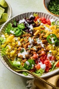 A colorful taco salad in a bowl with ground meat, black beans, corn, lettuce, diced tomatoes, shredded cheese, jalapeño slices, and a drizzle of creamy dressing. A fork rests on the side of the bowl.