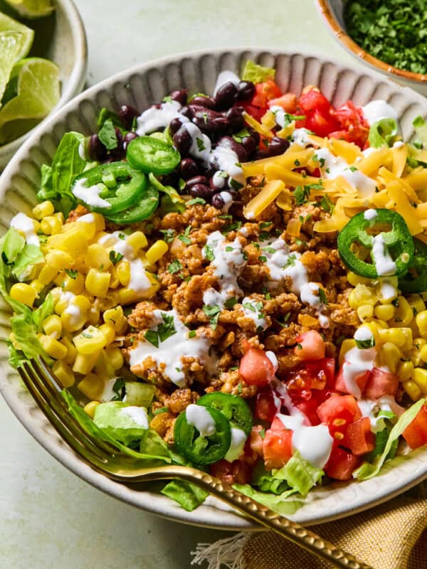 A colorful taco salad in a bowl with ground meat, black beans, corn, lettuce, diced tomatoes, shredded cheese, jalapeño slices, and a drizzle of creamy dressing. A fork rests on the side of the bowl.