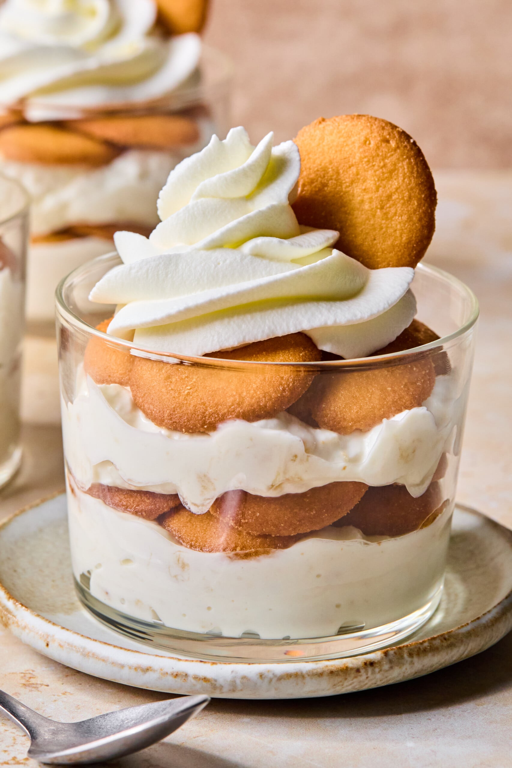 A glass dessert cup filled with layers of vanilla wafers and creamy pudding, topped with whipped cream and a vanilla wafer. The cup is placed on a small plate with a spoon nearby.
