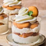 A glass dessert cup filled with layers of vanilla pudding, round cookies, and whipped cream, garnished with a cookie and a mint leaf, sits on a plate with a spoon beside it. Another dessert cup is in the background.