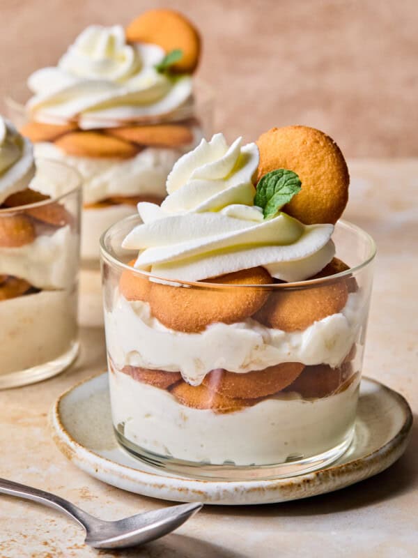 A glass dessert cup filled with layers of vanilla pudding, round cookies, and whipped cream, garnished with a cookie and a mint leaf, sits on a plate with a spoon beside it. Another dessert cup is in the background.