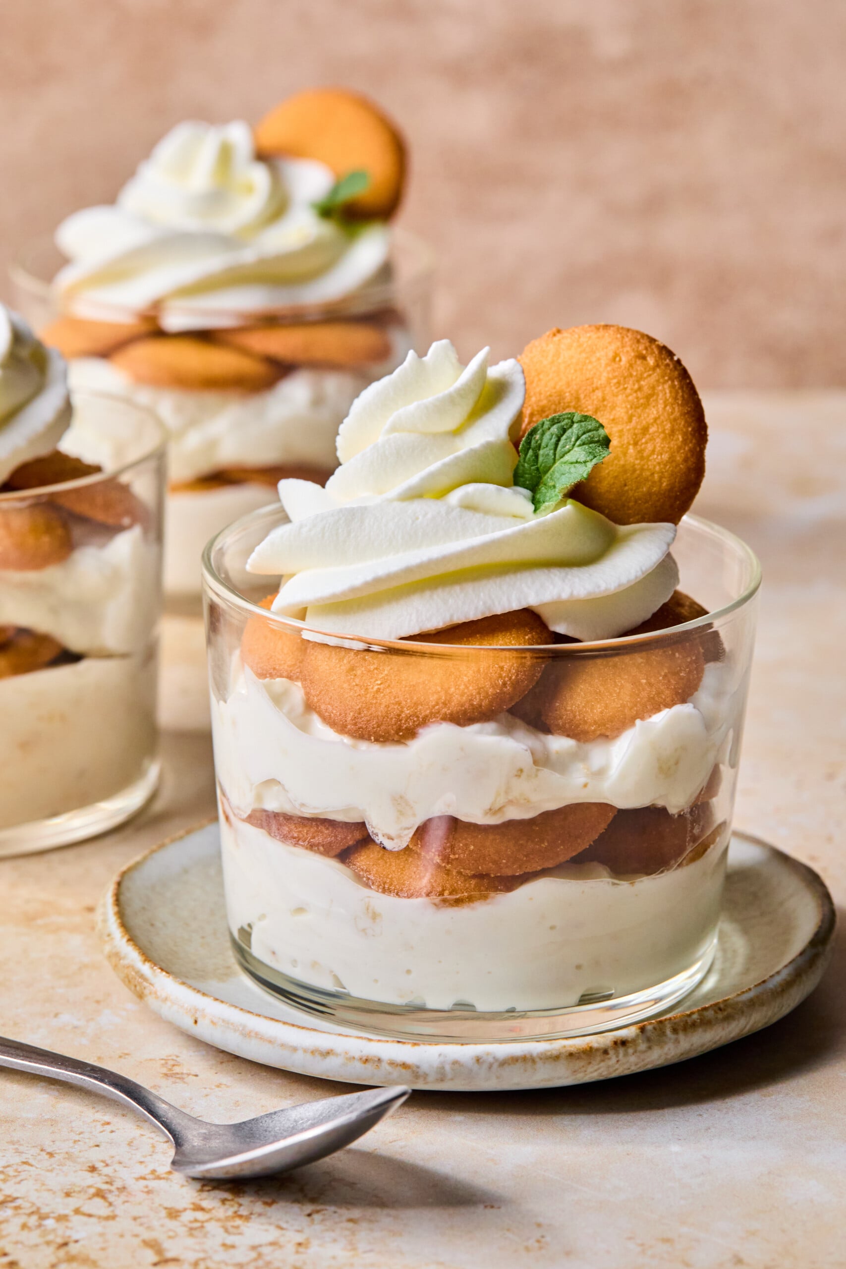 A glass dessert cup filled with layers of vanilla pudding, round cookies, and whipped cream, garnished with a cookie and a mint leaf, sits on a plate with a spoon beside it. Another dessert cup is in the background.