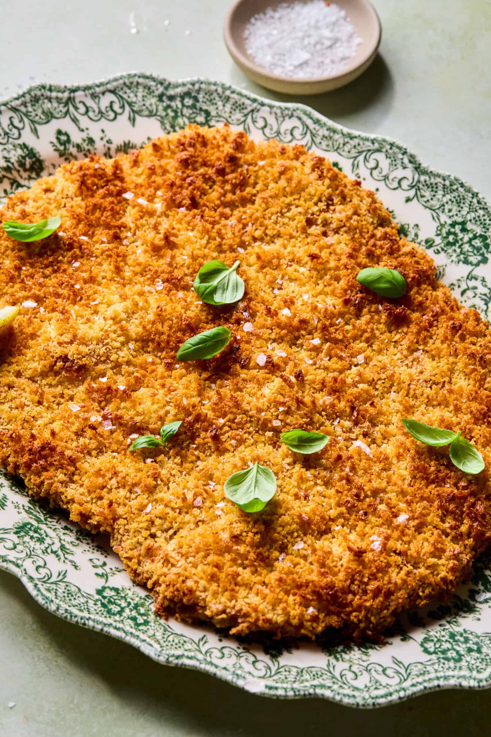 A large breaded and golden-brown schnitzel garnished with fresh basil leaves sits on a vintage green-and-white patterned platter, with a small dish of flaky salt in the background.