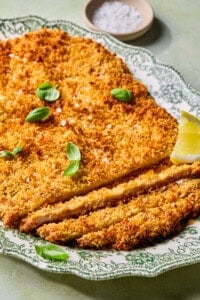A crispy breaded cutlet garnished with fresh basil leaves and coarse salt, served on an ornate green and white plate with a lemon wedge and a small dish of salt in the background.