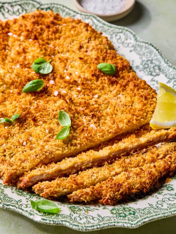 A crispy breaded cutlet garnished with fresh basil leaves and coarse salt, served on an ornate green and white plate with a lemon wedge and a small dish of salt in the background.