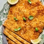 A large, breaded and baked schnitzel on a decorative plate, garnished with fresh basil leaves and lemon wedges, with a knife and several sliced pieces at one end. A small bowl of salt is in the background.