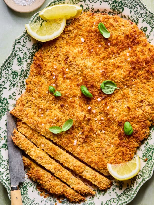 A large, breaded and baked schnitzel on a decorative plate, garnished with fresh basil leaves and lemon wedges, with a knife and several sliced pieces at one end. A small bowl of salt is in the background.