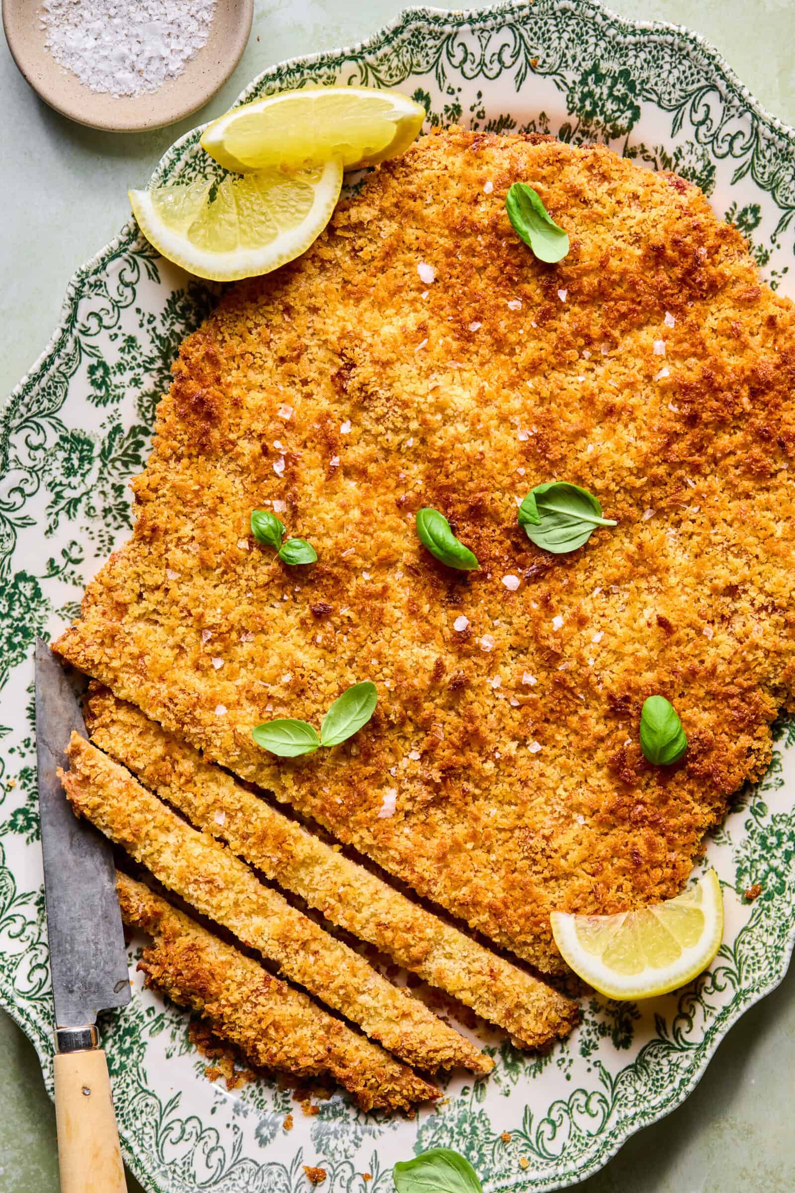 A large, breaded and baked schnitzel on a decorative plate, garnished with fresh basil leaves and lemon wedges, with a knife and several sliced pieces at one end. A small bowl of salt is in the background.