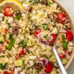 A close-up of a bowl of couscous salad with cherry tomatoes, cucumber, olives, red onion, feta cheese, and herbs. Above the bowl, a spoonful highlights fresh ingredients reminiscent of vibrant Italian eggplant parmesan flavors.