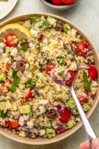 A close-up of a bowl of couscous salad with cherry tomatoes, cucumber, olives, red onion, feta cheese, and herbs. Above the bowl, a spoonful highlights fresh ingredients reminiscent of vibrant Italian eggplant parmesan flavors.