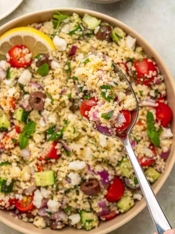 A close-up of a bowl of couscous salad with cherry tomatoes, cucumber, olives, red onion, feta cheese, and herbs. Above the bowl, a spoonful highlights fresh ingredients reminiscent of vibrant Italian eggplant parmesan flavors.