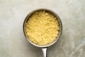 A top-down view of a saucepan filled with cooked, fluffy couscous on a light, textured surface.