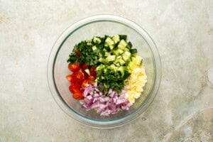 A glass bowl containing chopped red onion, cucumber, cherry tomatoes, cilantro, and crumbled cheese, arranged in sections on a light-colored countertop.