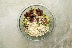 A glass bowl containing chopped olives, crumbled feta cheese, and a quinoa mixture with herbs and diced vegetables on a light, textured surface.