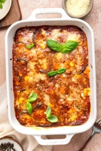 A baked eggplant parmesan casserole in a white dish, topped with melted cheese, tomato sauce, and fresh basil leaves, with a bowl of grated cheese nearby.