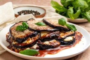 A plate of eggplant parmesan with layers of eggplant, tomato sauce, and melted cheese, garnished with fresh basil leaves. A bowl of pepper and fresh basil are visible in the background.