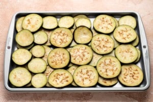 Slices of raw eggplant arranged in a single layer on a metal baking sheet, ready for roasting, on a light textured surface.