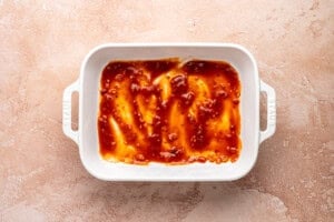 A white rectangular baking dish with handles contains a thin layer of reddish-brown sauce spread across the bottom, sitting on a textured light brown surface.