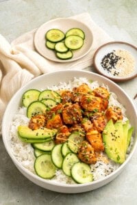 A bowl of white rice topped with glazed chicken pieces, sliced cucumber, avocado, and sprinkled with black sesame seeds, with extra cucumber slices and a dish of sesame seeds in the background.
