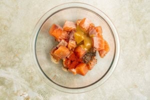 A glass bowl containing chunks of raw salmon mixed with mustard, ground spices, and seasoning on a light countertop.