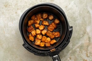 Chunks of cooked, seasoned chicken sit in the basket of an air fryer, resting on a textured, light-colored surface.