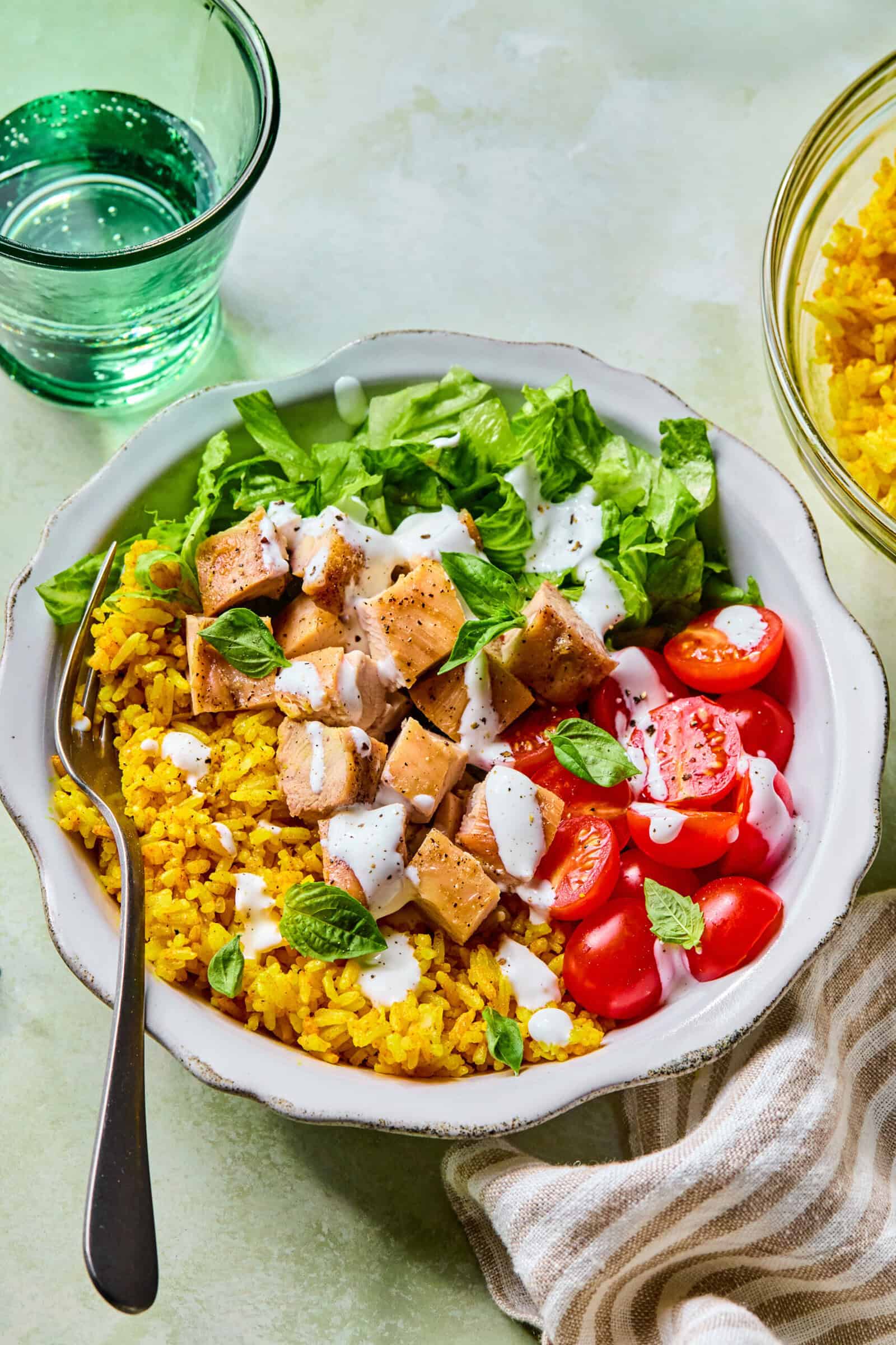 A bowl with yellow rice, chopped lettuce, diced chicken, halved cherry tomatoes, and fresh herbs, topped with a drizzle of white sauce. A fork rests in the bowl and a glass of water is beside it.