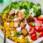A bowl with yellow rice, chopped grilled chicken, lettuce, halved cherry tomatoes, and fresh basil, topped with white dressing, with a fork on the side. A glass of water and a napkin are nearby.