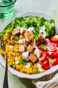 A bowl with yellow rice, chopped grilled chicken, lettuce, halved cherry tomatoes, and fresh basil, topped with white dressing, with a fork on the side. A glass of water and a napkin are nearby.
