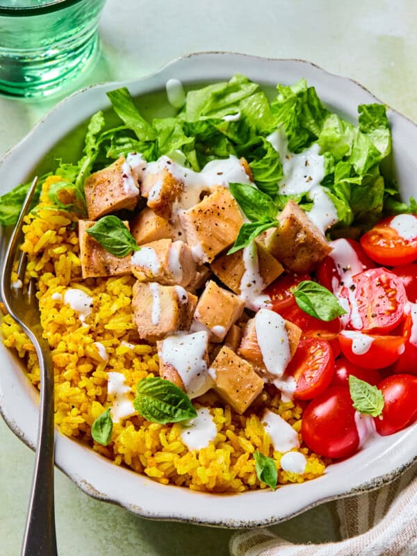 A bowl with yellow rice, chopped grilled chicken, lettuce, halved cherry tomatoes, and fresh basil, topped with white dressing, with a fork on the side. A glass of water and a napkin are nearby.