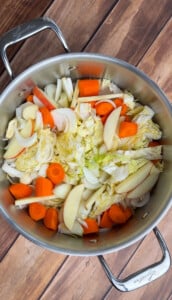 A stainless steel pot on a wooden surface contains chopped vegetables, including carrots, onions, napa cabbage, and apple slices—perfect ingredients for a flavorful no water chicken soup recipe, ready to be cooked.