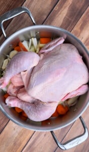 A whole raw chicken sits atop carrots, onions, and potatoes in a large stainless steel pot—perfect for preparing a flavorful no water chicken soup recipe on a rustic wooden surface.